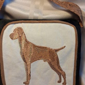 May include: A brown and black shoulder bag with a dog embroidery on the front. The bag has a brown strap with a paisley pattern and a black border around the dog image. The dog is brown and stands on a white background.