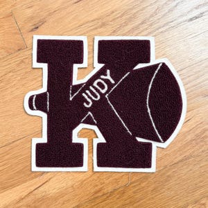 Könnte beinhalten: Ein weinrotes Letterman-Jacken-Patch in Form des Buchstabens "K" mit einem Megaphon-Design. Das Wort "JUDY" ist in Weiß auf dem Megaphon gestickt. Der Patch hat einen weißen Rand.