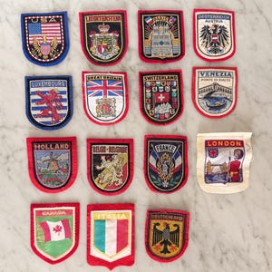 May include: A collection of fourteen embroidered souvenir patches featuring various national flags, city landmarks, and heraldic crests. Each shield-shaped patch displays colorful thread work, including text labels like USA, Paris, London, and Canada.