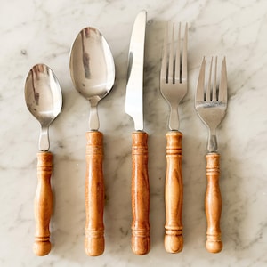 May include: A set of five pieces of flatware arranged in a row, consisting of a small spoon, a large spoon, a dinner knife, a dinner fork, and a small fork. Each piece features a polished stainless steel head and a rounded, light brown wooden handle.