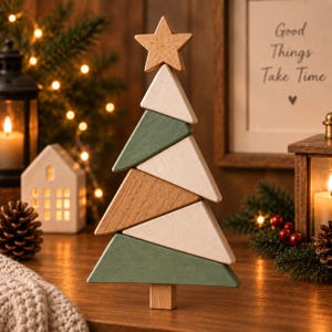 Puede incluir: Una escultura decorativa de árbol de Navidad de madera compuesta por segmentos triangulares apilados en tonos alternos de verde salvia, madera clara natural y blanco crema, rematada con una pequeña estrella de madera y una base rectangular.