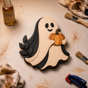 May include: A wooden ghost-shaped decoration featuring a light-coloured ghost holding an orange pumpkin with a dark brown stem. The ghost is layered over a dark black-stained wooden backing that includes a decorative leaf branch element.