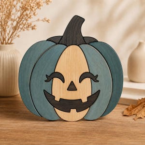 Peut inclure: Une décoration en bois en forme de citrouille avec un centre en bois clair sculpté d'un visage de jack-o'-lantern, incluant des yeux noirs avec des cils et un large sourire, entouré de segments en bois bleu sarcelle et d'une tige foncée.