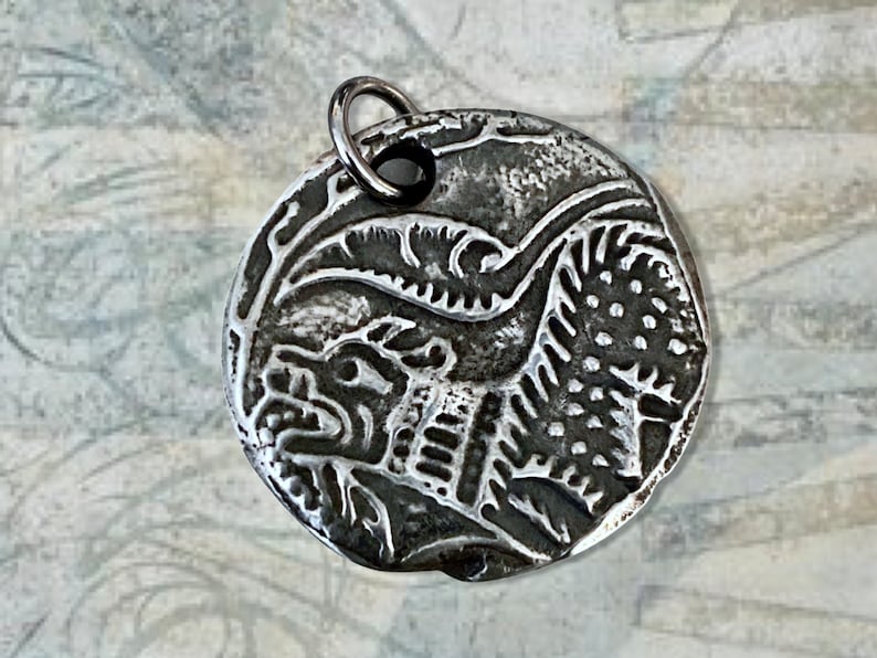May include: A silver pendant with a detailed design of a mythical creature with a horned head and a scaly body. The pendant is round and has a loop at the top for attaching a chain.