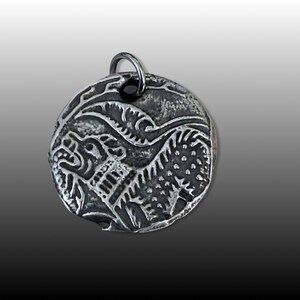 May include: Silver pendant with a detailed design of a mythical creature with a lion-like head and a body with scales. The pendant is round and has a loop at the top for attaching a chain.