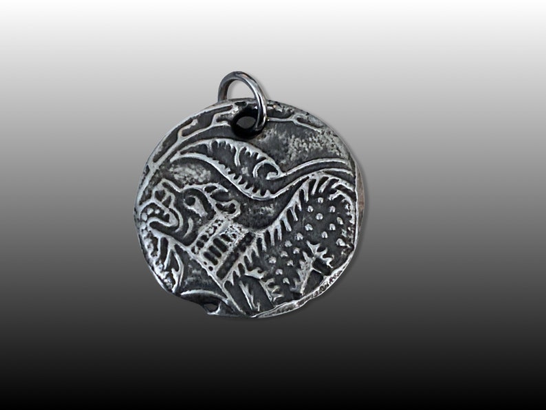 May include: Silver pendant with a detailed design of a mythical creature with a lion-like head and a body with scales. The pendant is round and has a loop at the top for attaching a chain.