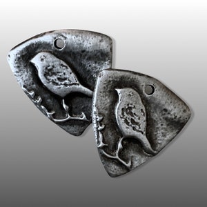 May include: Two silver metal pendants shaped like a bird on a branch. The pendants have a rustic, textured finish.