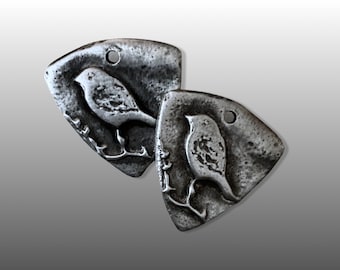 May include: Two silver metal pendants shaped like a bird on a branch. The pendants have a rustic, textured finish.