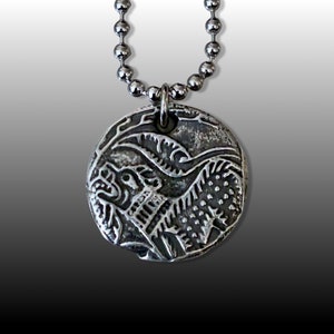 May include: A silver necklace with a round pendant featuring a detailed, embossed design of a mythical creature with sharp teeth and claws.