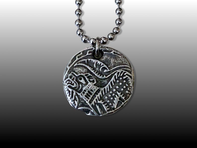May include: A silver necklace with a round pendant featuring a detailed, embossed design of a mythical creature with sharp teeth and claws.