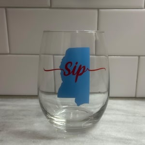 May include: A clear glass wine glass with a blue outline of the state of Mississippi and the word "Sip" in red cursive.