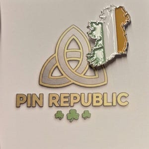 Puede incluir: Un pin metálico con el mapa de Irlanda con los colores de la bandera irlandesa. El pin está junto a un diseño de nudo celta dorado y plateado y las palabras "PIN REPUBLIC" con tres tréboles verdes.
