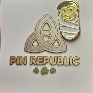 Pode incluir: Um alfinete de esmalte dourado com um bebê com auréola, envolto em um cobertor branco. O alfinete está em um fundo branco com um design de nó celta dourado e prateado e o texto "PIN REPUBLIC" com três trevos verdes.