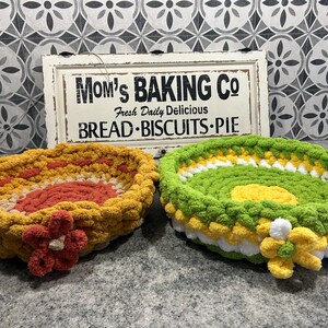 May include: Two handmade crochet baskets, one yellow and orange, the other green, yellow, and white. Each has a flower accent. A sign behind them reads "Mom's Baking Co" with text about bread, biscuits, and pie.