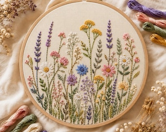 Op de afbeelding: Een ronde houten borduurring met een stoffen paneel met een gedetailleerd handgestikt ontwerp van diverse wilde bloemen in tinten geel, roze, blauw, paars en groen, vergezeld door verschillende losse strengen gekleurd borduurgaren.
