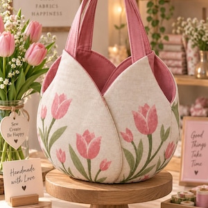 May include: A fabric tote bag shaped like a tulip flower, featuring a cream-colored exterior with a pink and green tulip print. The bag has two pink handles and a solid pink interior lining, designed with overlapping petal-like panels.
