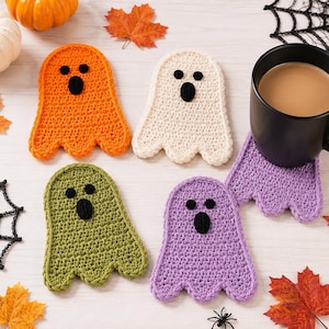 May include: A set of four ghost-shaped crochet coasters in orange, cream, olive green, and purple, each featuring two small black circular eyes and an oval black mouth. One purple coaster supports a black ceramic mug filled with coffee.