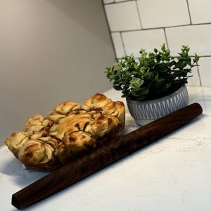 May include: A dark brown wooden rolling pin lies on a white surface beside a baked pastry and a small potted plant. The rolling pin is long and rectangular. The pastry is golden brown and swirled. The plant is green with a gray and white pot.