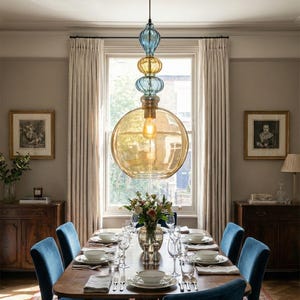 May include: A dining room with a large, multi-tiered glass pendant light fixture. The light fixture has a large amber-colored globe at the bottom, with blue and yellow glass spheres above. A long wooden dining table is set with plates, silverware, and glasses, with blue velvet chairs.