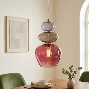 May include: A pendant light fixture featuring three stacked, translucent glass globes in shades of light purple, amber, and deep rose. The glass sections are separated by brass-toned metal hardware, with a single exposed filament bulb inside.