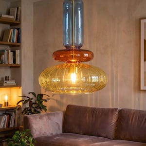 Könnte beinhalten: Moderne Pendelleuchte mit mehreren Ebenen. Der obere Teil ist ein blauer Glaszylinder, gefolgt von einer orangefarbenen Scheibe und einem großen, gerippten gelben Glasschirm. Die Leuchte hängt an einer goldfarbenen Stange.