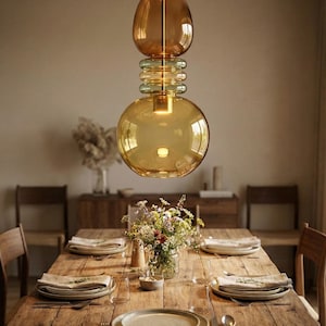 May include: A modern amber glass pendant light hangs above a rustic wooden dining table set for a meal. The light fixture has a bulbous shape with a large globe at the bottom and a smaller, egg-shaped top. The table is set with plates, silverware, and a vase of flowers.