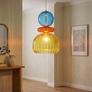 Könnte beinhalten: Eine moderne Pendelleuchte mit einem geschichteten Design. Sie verfügt über eine blaue Kugel, eine orangefarbene Scheibe und einen gerippten gelben Glasschirm. Die Leuchte hängt an einem dünnen Draht und erhellt den Raum mit einem warmen Glanz.