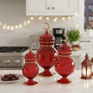 Könnte beinhalten: Drei rote dekorative Glasvasen mit goldenen Akzenten und einem Halbmond-Aufsatz. Die unterschiedlich großen Vasen stehen auf einer weißen Arbeitsplatte. Ein Teller mit Datteln und eine Laterne sind ebenfalls zu sehen.