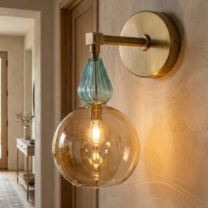 May include: A wall sconce featuring a circular brushed brass backplate and a horizontal arm. The fixture includes a ribbed, teardrop-shaped light blue glass element positioned above a large, spherical, amber-tinted glass shade with a light bulb.