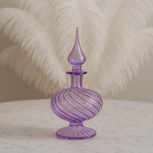 Può includere: Un flacone di profumo decorativo in vetro color lavanda con un design a spirale. Il flacone ha un corpo bulboso, un collo sottile e un tappo a punta. Si trova su una base rotonda ed è esposto su un tavolo di marmo bianco.