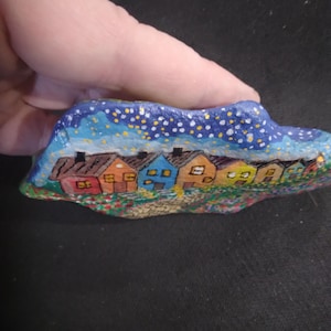 Può includere: Arte rupestre dipinta a mano con una fila di case colorate sotto un cielo stellato. Le case hanno tetti rossi, arancioni, blu e gialli. L'opera è tenuta in mano.