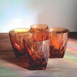 May include: Four amber-colored drinking glasses with a textured, swirling design. The glasses have a square base and a slightly flared rim. The glasses are arranged on a wooden surface, with a soft light in the background.