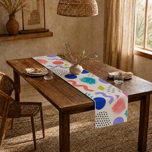May include: A white table runner featuring a colourful abstract pattern of blue, pink, and green geometric shapes, including circles, wavy lines, and polka dots, laid across a dark brown wooden dining table with two place settings and a vase.