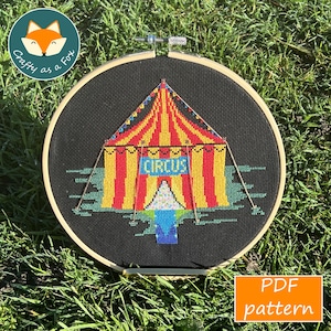 May include: Cross-stitch embroidery of a circus tent with red and yellow stripes, set against a black background. The word "CIRCUS" is displayed on a blue sign. The design is framed in a wooden hoop, with a "Crafty as a Fox" logo. A PDF pattern is available.