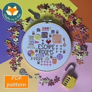 May include: A circular embroidery hoop containing a cross-stitch pattern with the text I ESCAPE ROOMS surrounded by icons of keys, locks, a maze, a clock, and lightbulbs, accompanied by scattered puzzle pieces, two metal keys, and a yellow padlock.