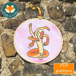 Peut inclure: Un cercle de broderie au point de croix avec un motif abstrait orange et jaune sur fond rose. Le cercle en bois est suspendu à un ruban jaune. Le texte "PDF pattern" est dans un encadré.