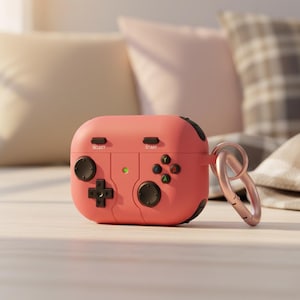 May include: A coral-colored AirPod case designed to resemble a video game controller. The case features black buttons, a directional pad, and the words "SELECT" and "START". A rose gold carabiner clip is attached to the side.