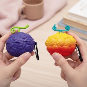May include: Two colorful, fruit-shaped AirPod cases. One is purple with a green stem, the other is yellow to red with a blue stem. Both cases have a textured surface and black carabiner clips.
