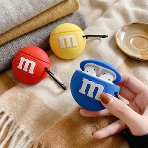 Puede incluir: Tres fundas de silicona coloridas para AirPods en rojo, amarillo y azul, cada una con un logotipo "M" blanco. La funda azul está abierta, revelando auriculares blancos. Cada funda tiene un mosquetón negro para sujetar.