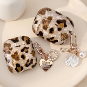 Peut inclure: Deux étuis pour AirPods avec un motif léopard crème, marron et noir. Un étui est ouvert et l'autre est fermé. Chaque étui a un porte-clés doré avec un charme. Un charme est un cœur avec du texte, l'autre est un coquillage.