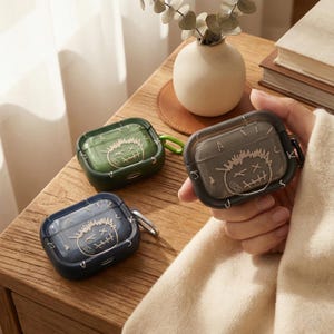 May include: Three protective cases for wireless earbuds in green, navy blue, and gray. Each case features a unique graphic design with the text "JACKS" and a cartoon face. The cases have a carabiner clip for easy attachment.