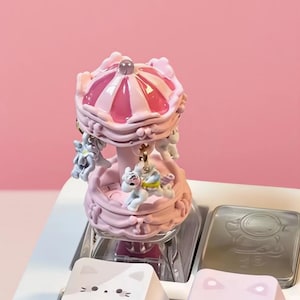 May include: A pink and white carousel-themed keyboard keycap. The keycap features a miniature carousel with small unicorn figures and a striped roof. The keycap is part of a custom keyboard setup with other cat-themed keycaps.