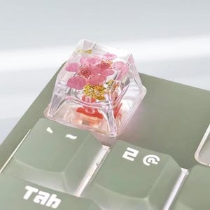 Può includere: Un copritasto trasparente quadrato con fiori rosa e scaglie d'oro. Il copritasto è posizionato su una tastiera verde, con il tasto "Tab" visibile.