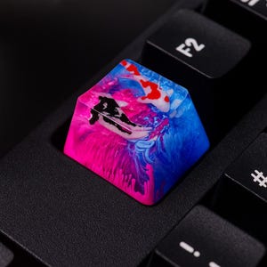 Puede incluir: Una tecla de teclado personalizada con un diseño de carpa koi. La tecla es una mezcla de resina rosa, azul, blanca y roja, con una silueta negra de carpa koi. La tecla está instalada en un teclado negro.