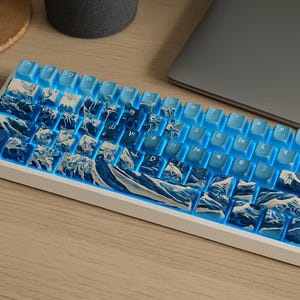 Op de afbeelding: Een witte mechanische toetsenbord met blauwe toetsen met een berg- en golfontwerp. Het toetsenbord heeft een blauwe achtergrondverlichting en staat naast een laptop op een houten oppervlak.
