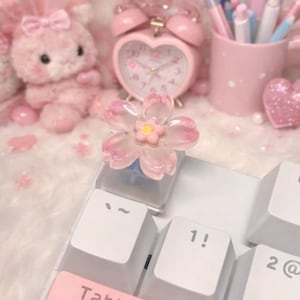 Può includere: Un copritasto a forma di fiore rosa è posizionato su una tastiera bianca. Il copritasto è circondato da accessori rosa, tra cui una sveglia a forma di cuore, un coniglietto di peluche e un portapenne. La tastiera ha il tasto 1.