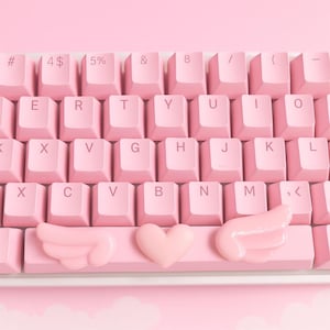 Pode incluir: Um teclado mecânico rosa com teclas rosa claro. O teclado apresenta uma barra de espaço personalizada com um design de coração e asas. As teclas estão dispostas em um layout QWERTY padrão.