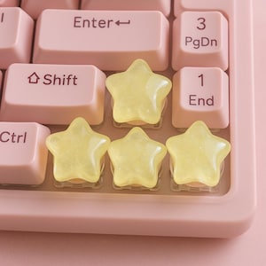 Może przedstawiać: Zbliżenie różowej klawiatury komputera z żółtymi obiektami w kształcie gwiazd. Klawisze klawiatury są jasnoróżowe, widoczne klawisze "Enter", "Shift", "Ctrl", "1", "3" i "End". Obiekty w kształcie gwiazd są przezroczyste i żółte.