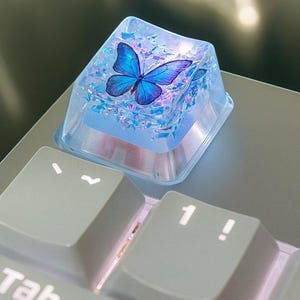 Op de afbeelding: Een aangepaste toetsenbordtoets met een blauw vlinderontwerp in heldere hars. De vierkante toets zit op een lichtgrijs toetsenbord. De vlinder heeft blauwe en paarse vleugels, een uniek accessoire.