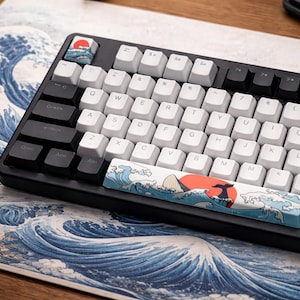 Puede incluir: Un teclado mecánico negro con teclas blancas y negras. La barra espaciadora y una tecla de escape presentan un diseño de olas japonesas con un sol rojo. El teclado descansa sobre una alfombrilla de ratón a juego con un patrón de olas similar.